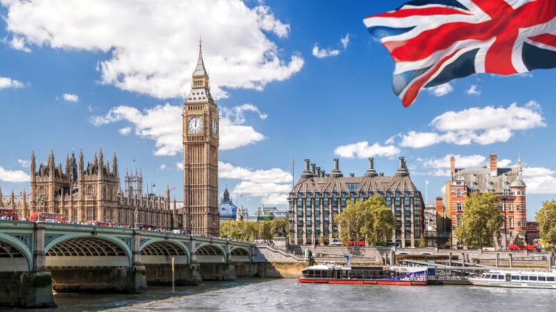 Big Ben with bridge over Thames and flag of England against blue sky in London